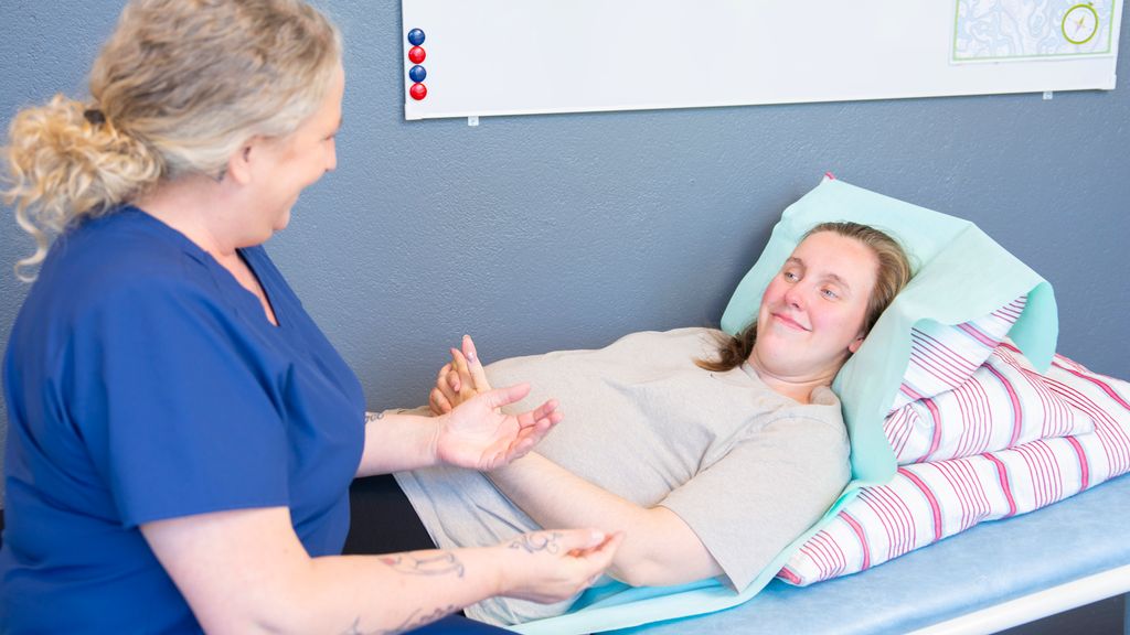 Gravid kvinne ligg på ein undersøkingsbenk og har hendene på magen. Ein person i uniform for helsepersonell sit på kanten av undersøkingsbenken. Dei har augekontakt og smiler til kvarandre. Foto.