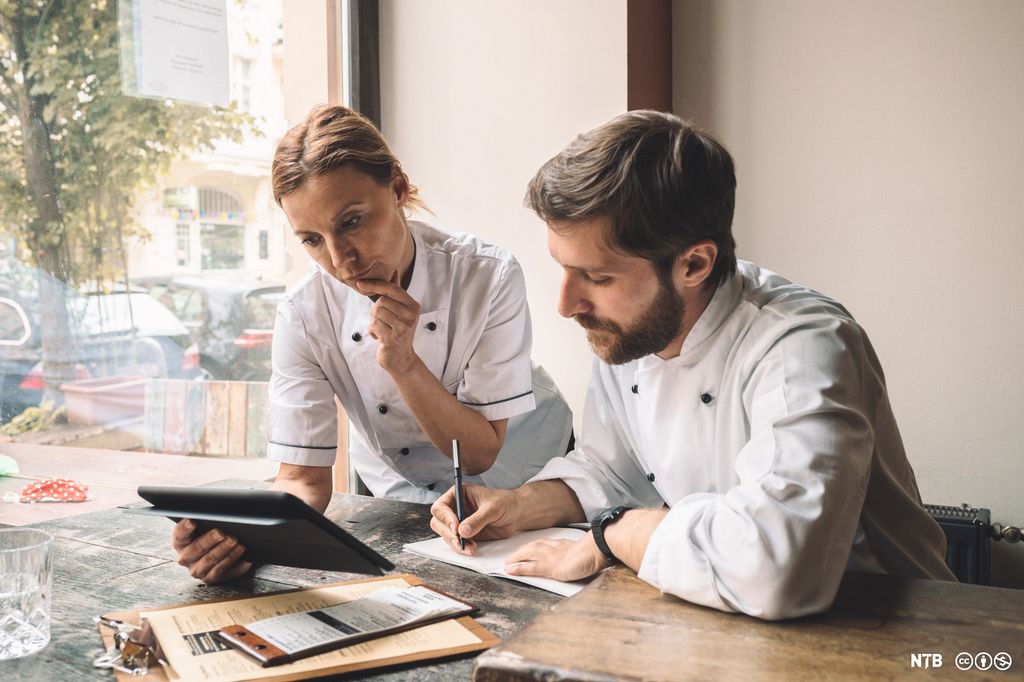 To kokker ser på et nettbrett mens de skriver i ei notatbok. Foto.