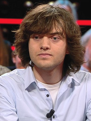 Portrait of Boyan Slat. He has shoulder length brown hair, is wearing a light blue shirt. Photo.