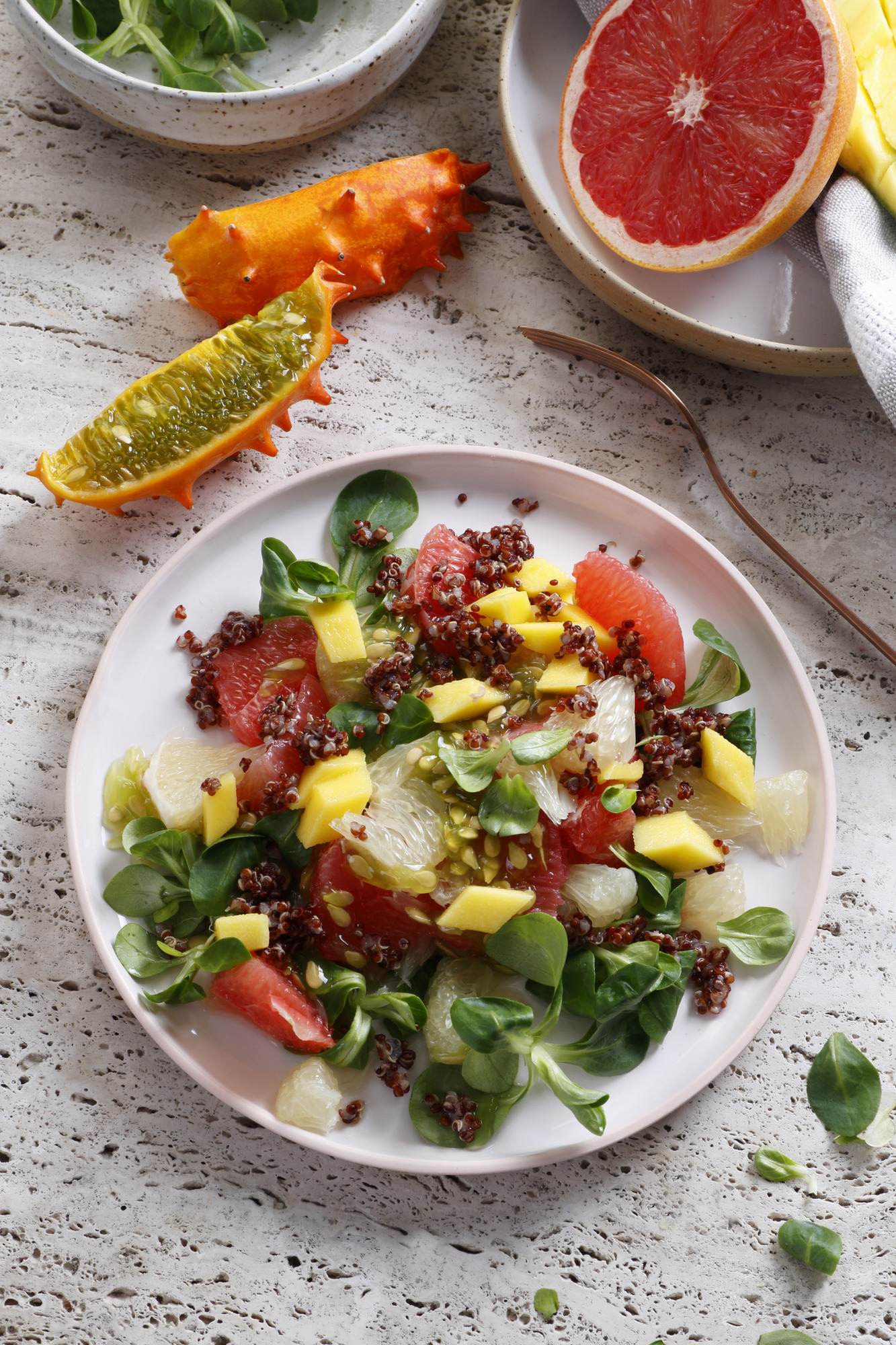 Fargerik salat anrettet på hvit tallerken. På bordet ved siden av tallerkenen ligger det en delt kiwano og en delt grapefrukt. Foto.