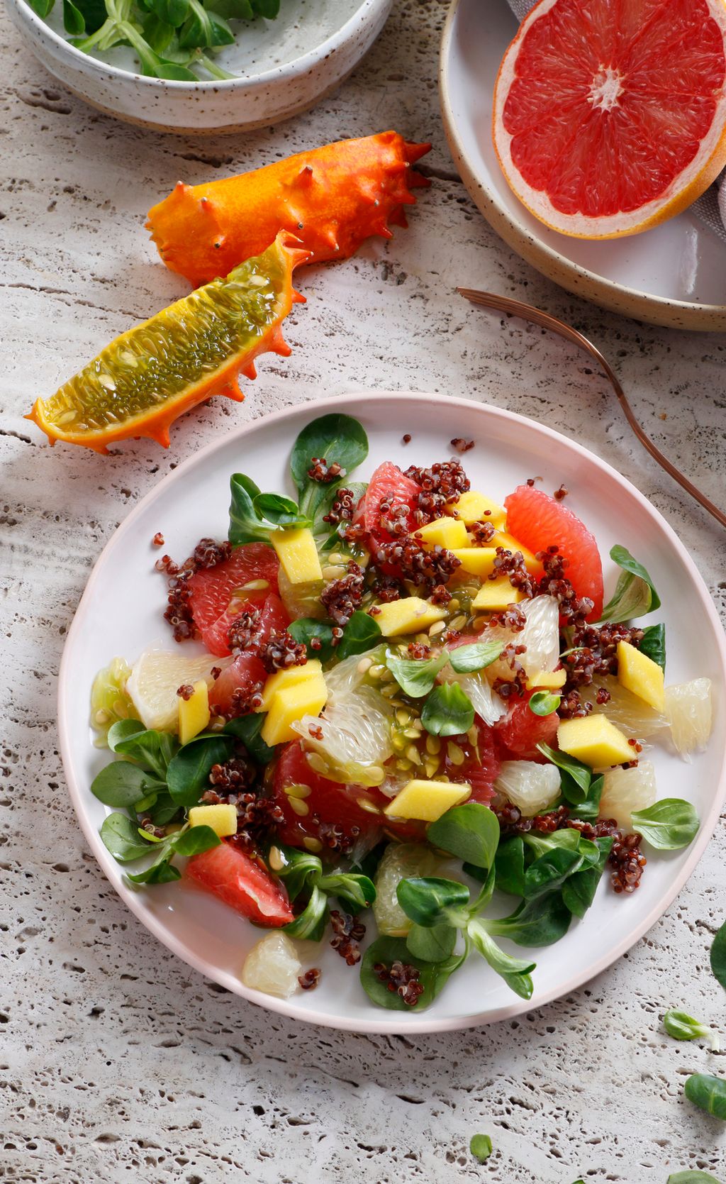 Fargerik salat anrettet på hvit tallerken. På bordet ved siden av tallerkenen ligger det en delt kiwano og en delt grapefrukt. Foto.