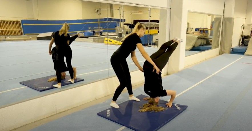 Jente som prøver å stå på hodet, og ei annen jente som støtter henne og holder hendene på hoftene hennes. Foto.