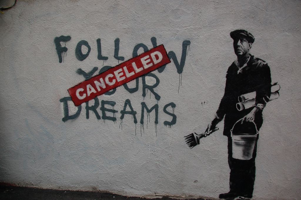 Graffiti i svart og hvitt på murvegg av en som arbeider med å klistre opp plakater. Ved siden av mannen står det skrevet "Follow your dreams". Over teksten er det skrevet "Cancelled" i hvite bokstaver på rød bakgrunn. Foto.