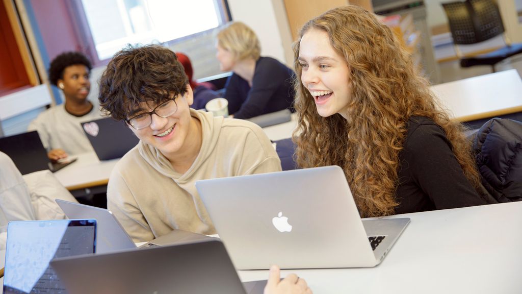 Elever sitter ved pultene sine og diskuterer. De har bærbare laptopper foran seg. Foto.