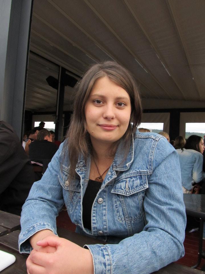 Smilende ung kvinne sittende ved et utebord. Hun har langt, mellombrunt hår og brune øyne, og hun har på seg lyseblå olajakke. Foto.