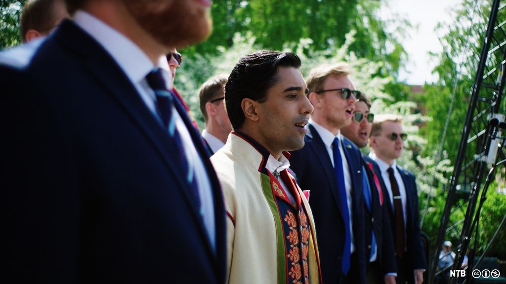 En mørkhudet ung mann i bunad står og synger sammen med andre unge menn i dress.  Foto.