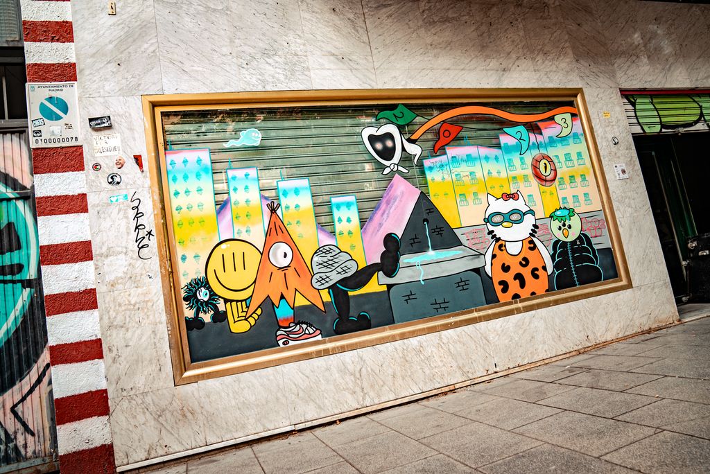 Vegg med et metallgitter foran et stort vindu. Glasset foran gitteret er dekorert med et stort maleri som viser høye skyskrapere i lyse farger i bakgrunnen, tegneserieliknende figurer i forgrunnen. Foto.