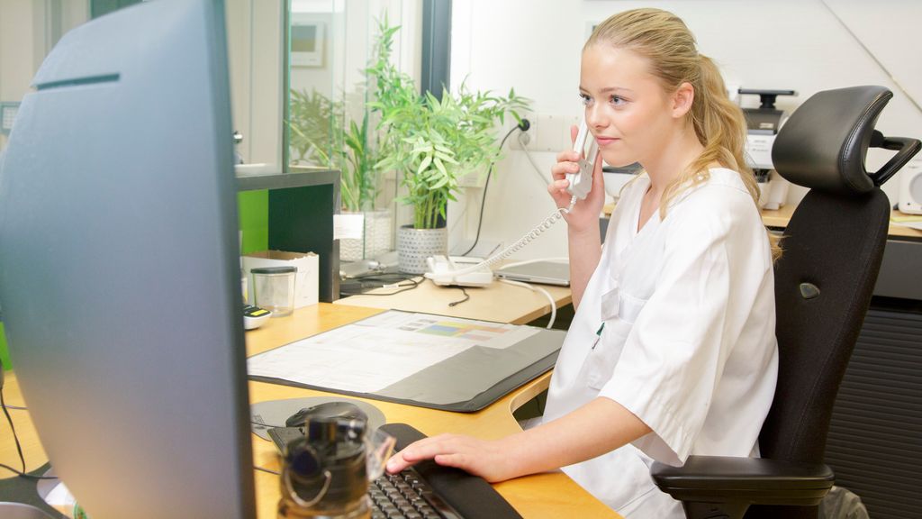 Helseservicearbeidar sit framfor pc i resepsjonen og snakkar i telefonen. Foto.