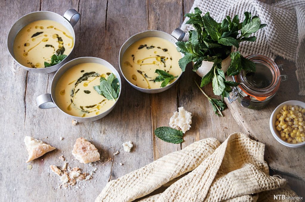Tre skåler kremet, lys suppe, sammen med brødbiter og mais på et bord. Foto.