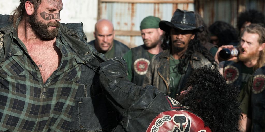 Photo: We see a man with a tattooed face fighting another man who is grabbing on to his shoulder. A group of men are waching in the background. 