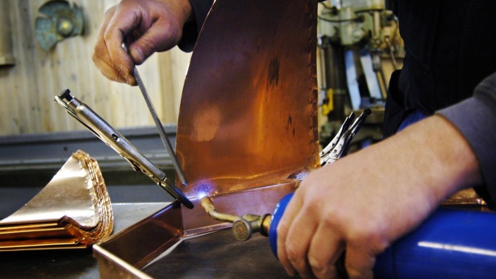 Loddebolt varmes opp med propanflamme. Ei kobberplate blir sveiset fast. Flere buede kobberplater ligger ved siden av. Foto.