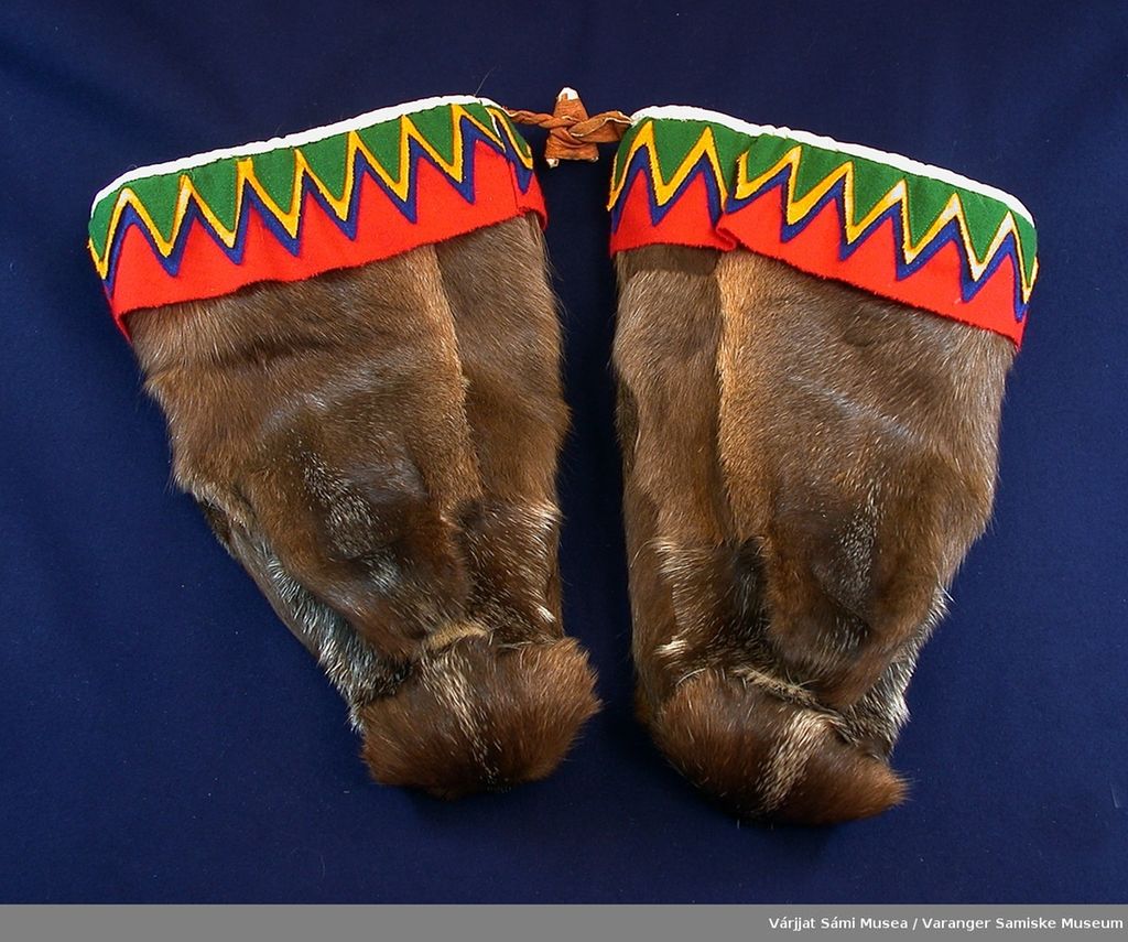 Fotografi av et par pelsvotter. I åpningen rundt håndleddet har de et bredt pyntebånd i tekstil med sikksakkmønster i fargene rødt, blått, gult og grønt.