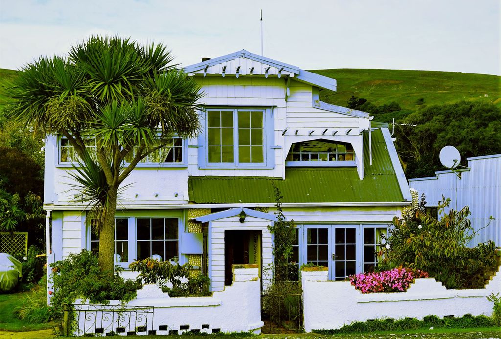 A white house with a green roof, blue details, and a white garden fence. It is surrounded by green nature. Photo.