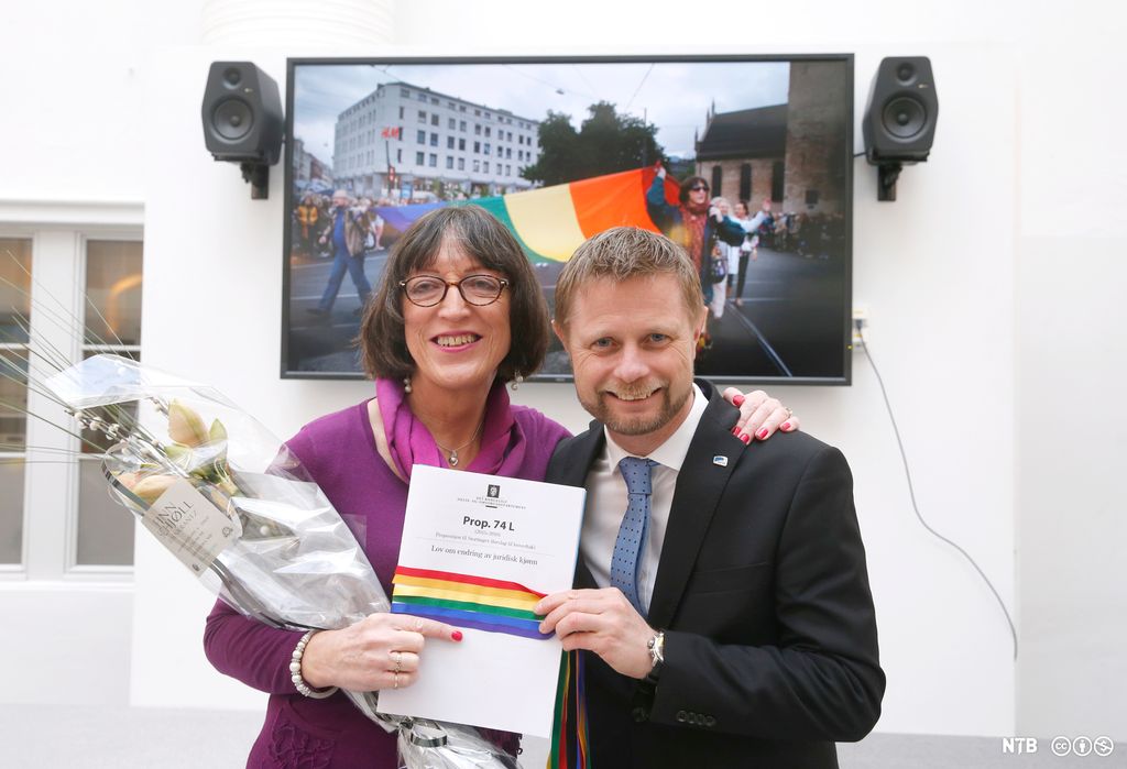 En kvinne og en mann holder et dokument med regnbuefarget bånd. Foto.