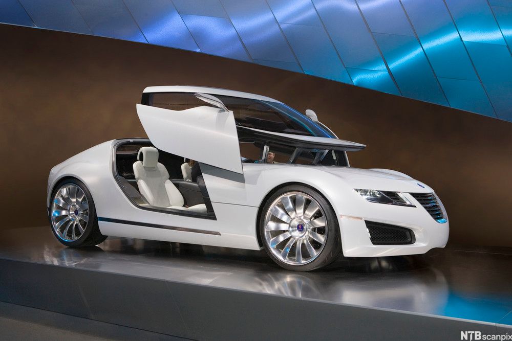 En stilig hvit bil med hevedører står på utstilling på en plattform. Foto.