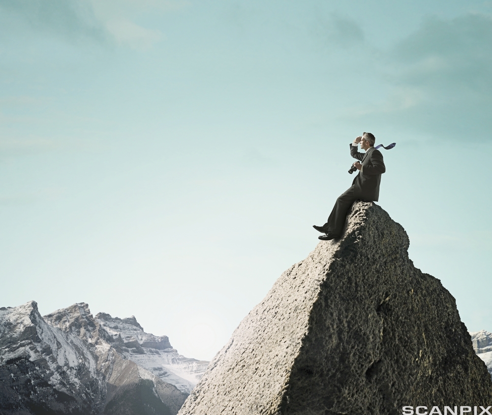 En dresskledd mann på toppen av et fjell holder en kikkert og speider utover. Foto. 