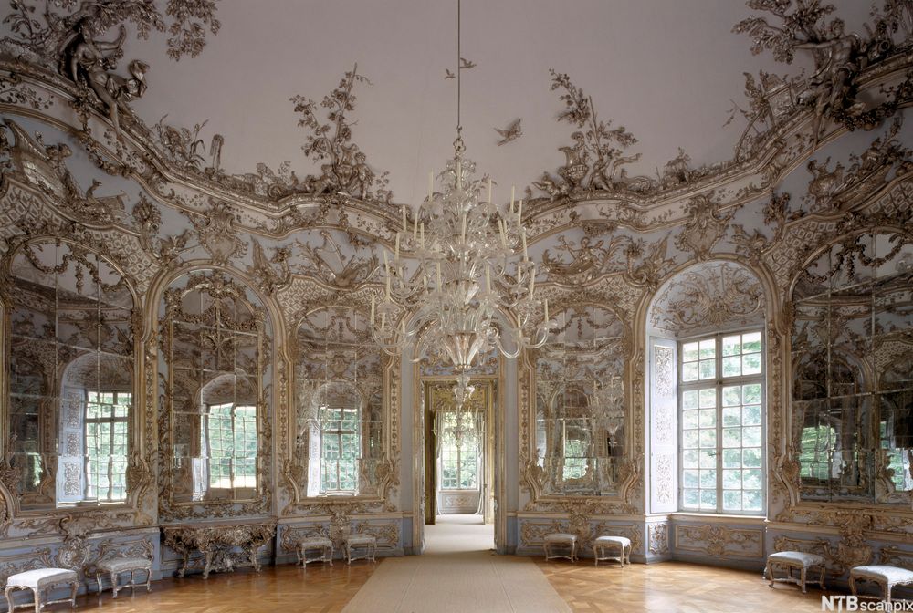 En hall med store speil og vinduer ut mot naturen. Det står skamler ved vinduene. Veggene er utsmykket med ornamenter i gullfarge. Foto.