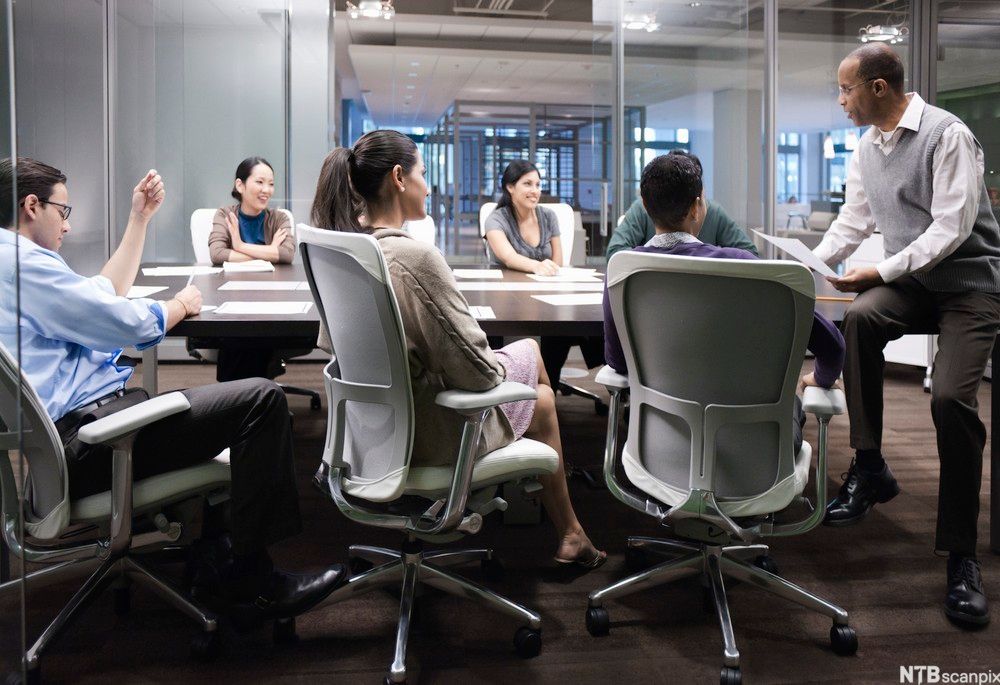 Seks personer sitter i hver sin stol rundt et møtebord og lytter til en mann som sitter på bordhjørnet og snakker. Foto.