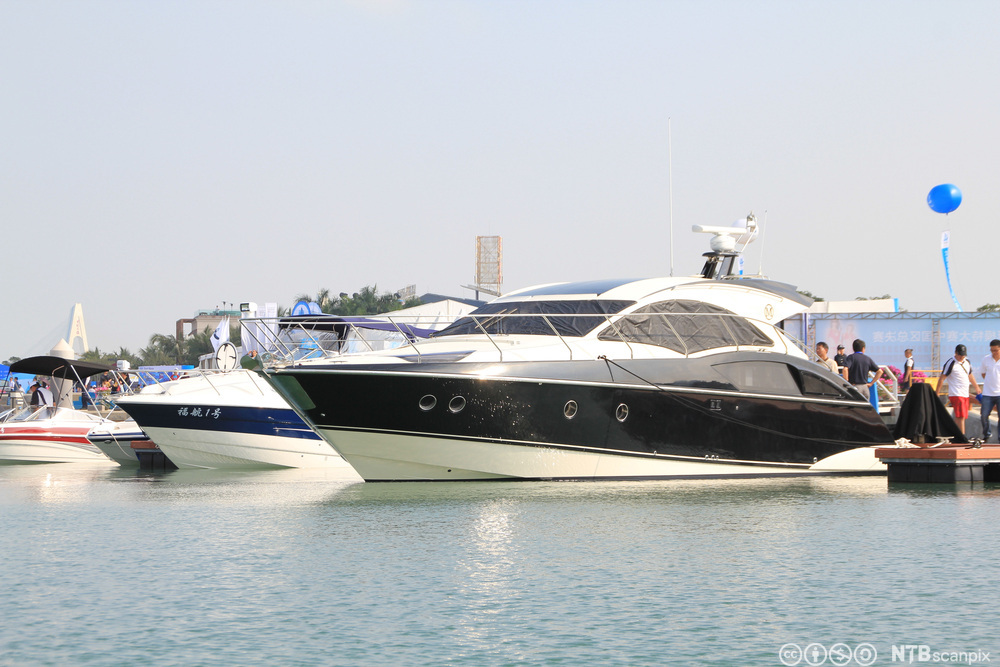 Luksuriøs sort og hvit cabincruiser ligger ankret opp ved siden av tre andre mindre båter. Det står noen personer på kaien. Foto.