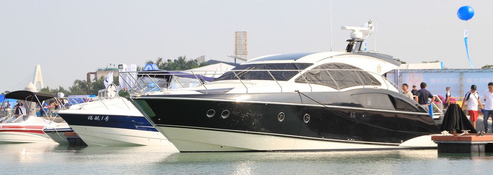 Luksuriøs sort og hvit cabincruiser ligger ankret opp ved siden av tre andre mindre båter. Det står noen personer på kaien. Foto.