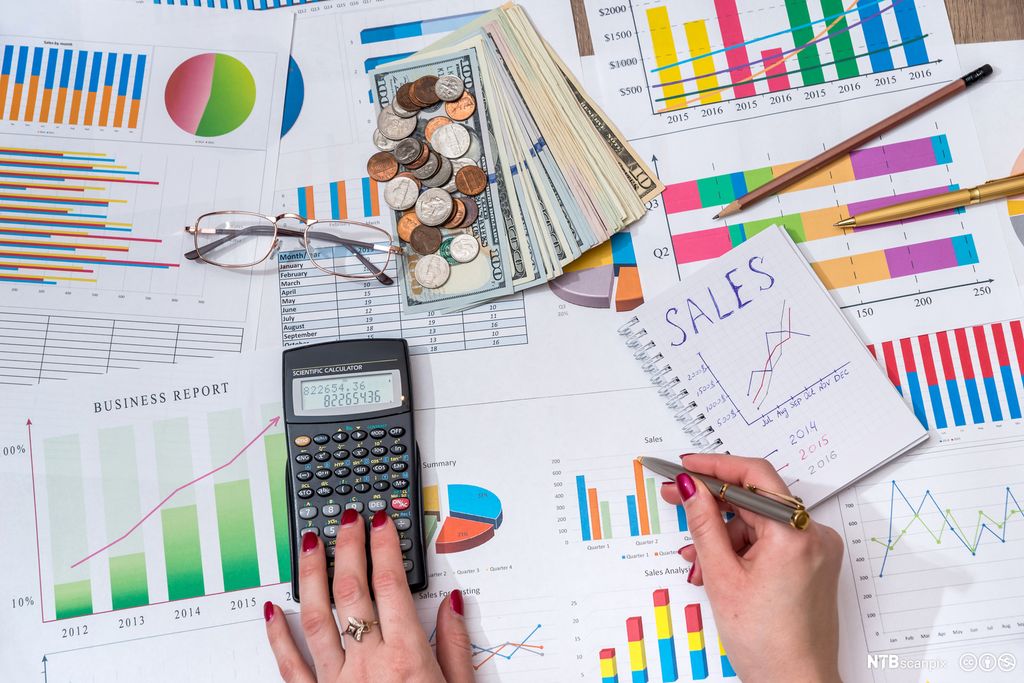 To kvinnehender på et bord med kalkulator, penn, notatblokk og ulike grafer. Foto.