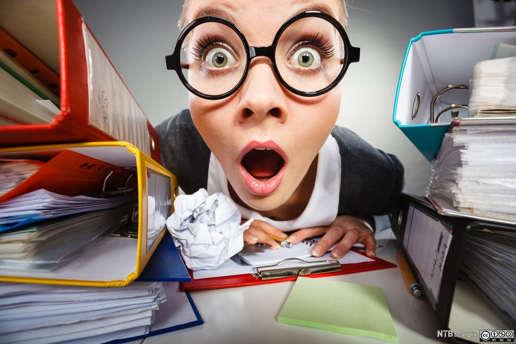 Ein rekneskapsmedarbeidar med briller ser overraska mot kameraet med open munn. På bordet framfor henne er det eit oppkrølla ark og ei notatbok. På begge sidene av bordet er det fleire dokumentmapper. Foto.