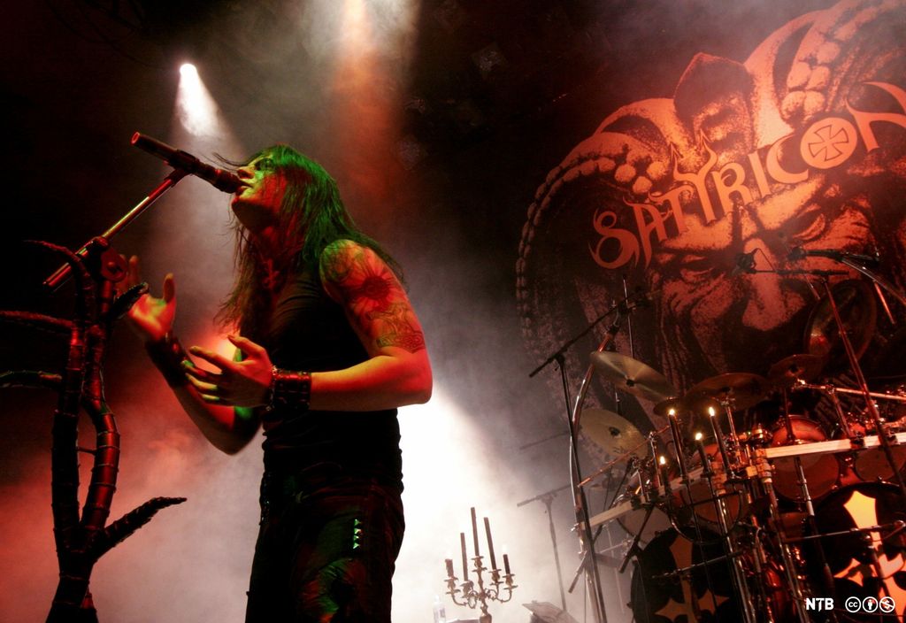 Vokalisten i Satyricon på scenen. Han har langt hår, svart singlet og svart bukse med nagler, naglearmbånd, tatovert overarm og et kors som henger opp ned rundt halsen. I bakgrunnen er det kandelabre, og veggen bak scenen er dekorert med et stort djevelansikt og logoen til Satyricon. Bokstaven o i logoen inneholder et korssymbol, og bokstaven y ser ut som en totannet djevelgaffel. Foto.