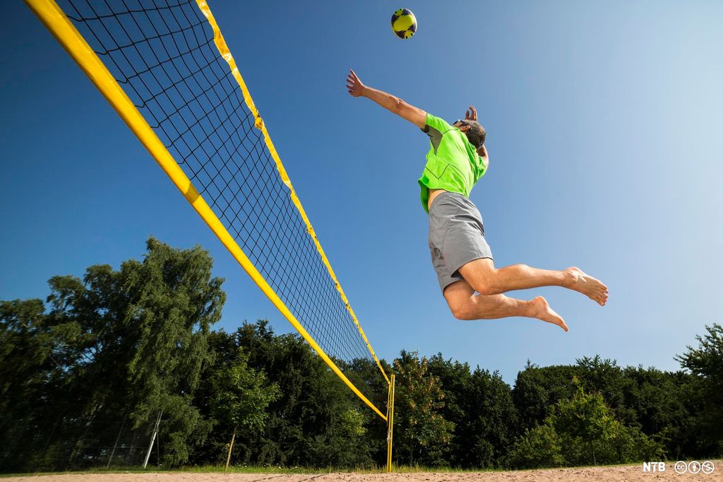 Foto av mann i grå shorts og grønn trøye som svever i lufta og er klar til å smashe en volleyball. I bakgrunnen er det grønn løvskog og blå himmel.