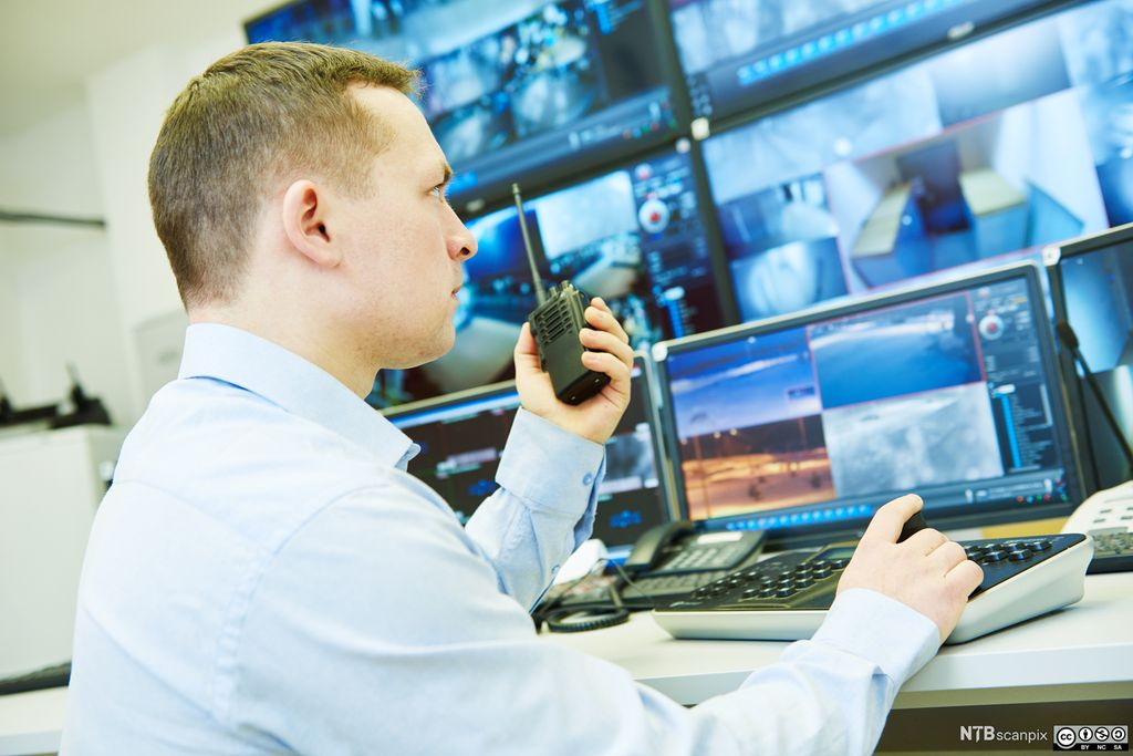 Sikkerhetsvakt sitter foran mange skjermer med video fra overvåkingskameraer. Foto.