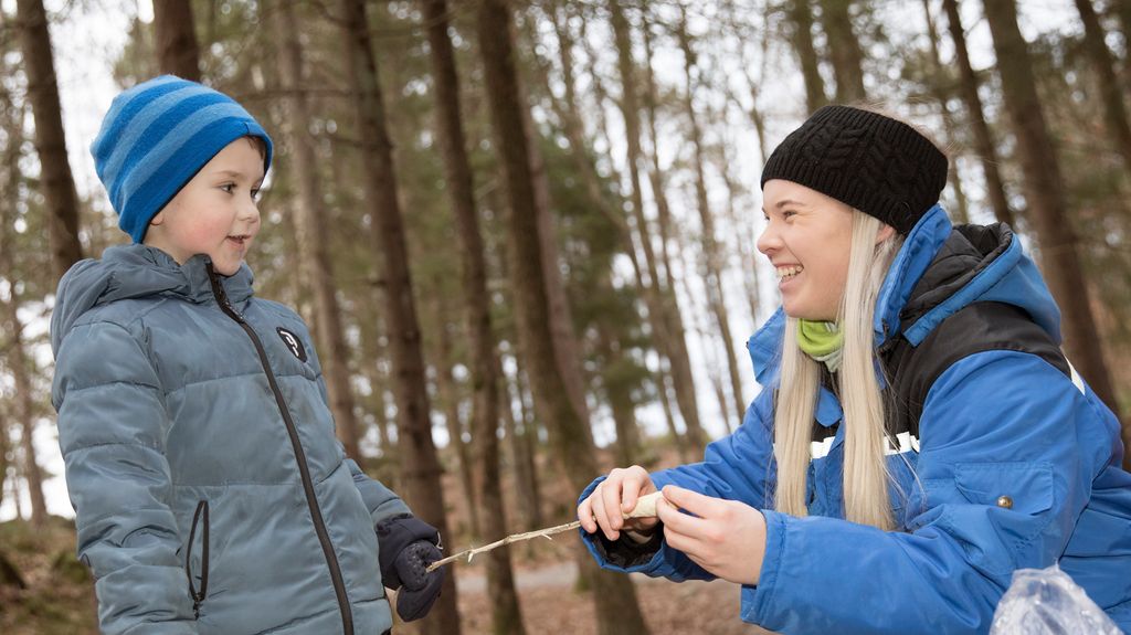 Gutt med vintertøy får hjelp av en voksen kvinne til å tvinne pinnebrød på en pinne. De smiler til hverandre. Foto.