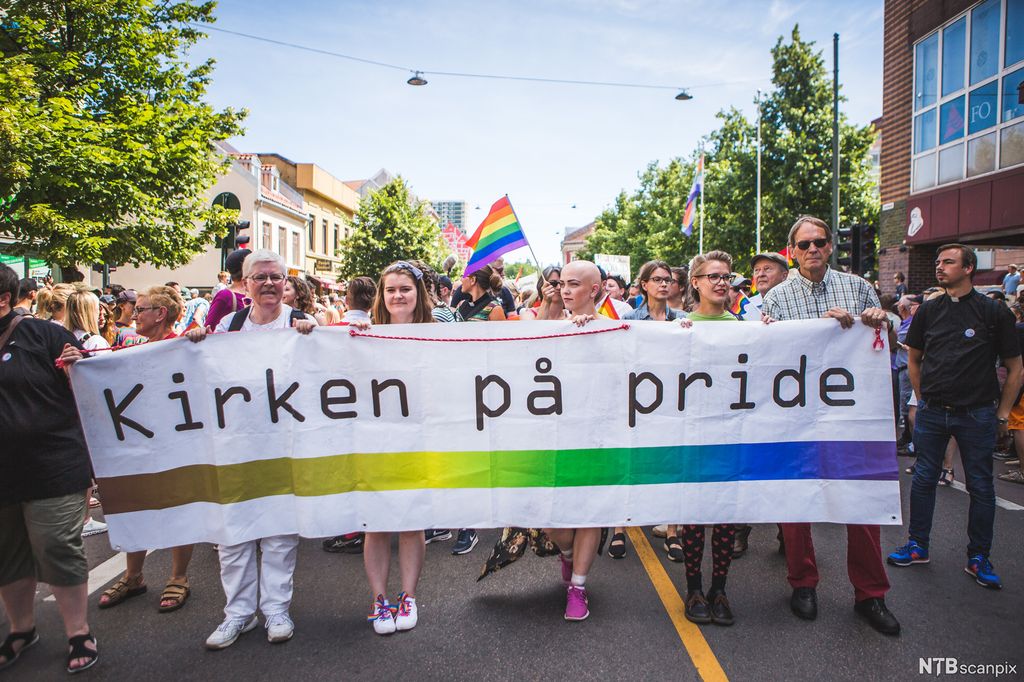 Fem personer bærer et banner med påskriften "Kirken på pride". Bak vaier regnbueflagget, og det er mange personer som går i en parade. Foto.