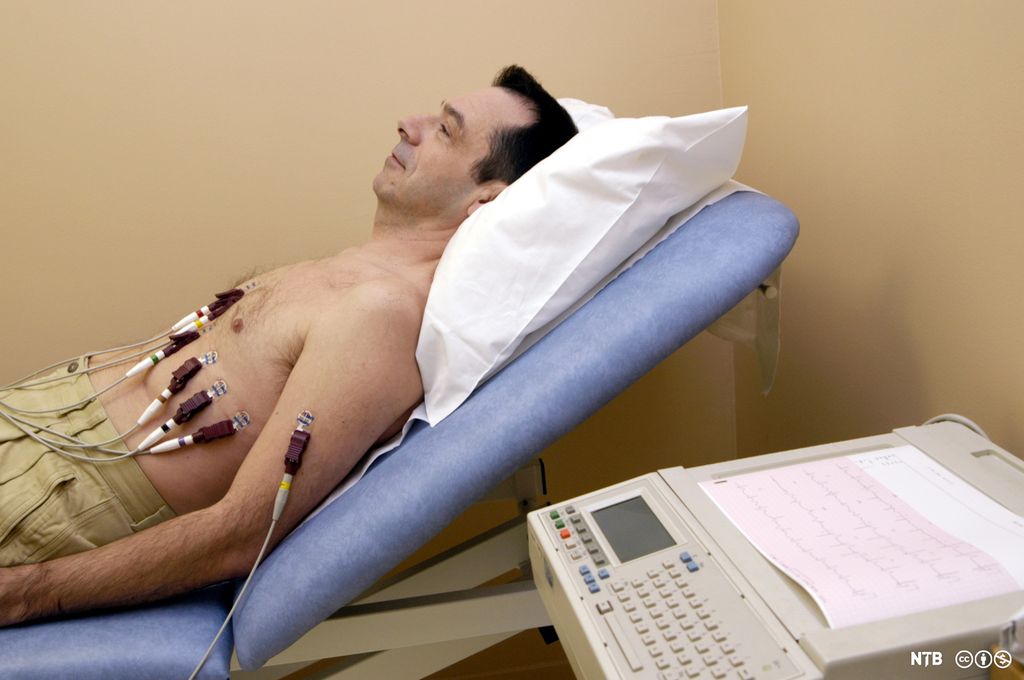 Mann i bar overkropp ligger på undersøkelsesbenk koblet til elektroder fra EKG-apparat. Foto.