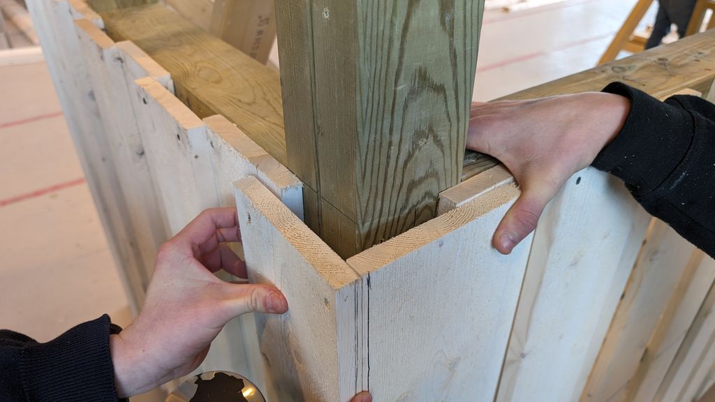 Hjørne med tømmermannskledning. To personer holder overliggerne som skal brukes på hjørnet, på plass der de skal monteres. Overliggeren på venstre side er trukket så langt ut at den dekker enden på overliggeren til høyre. Foto.