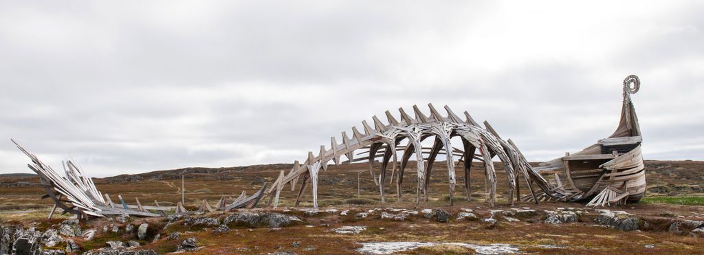 Treskulptur ute i et viddelandskap. Formen minner om baugen på et vikingskip som går over i et dyreskjelett og ender opp i en hale. Foto.