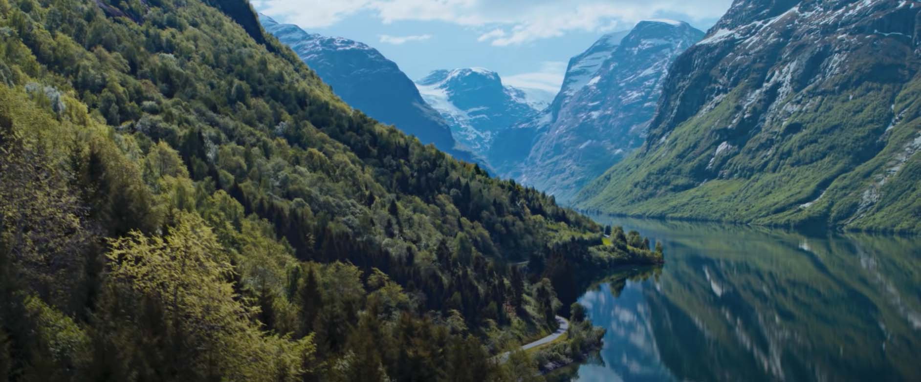 Bratte, grønne fjellsider som speiler seg i en blank fjord. Høye fjell med snø i bakgrunnen. Foto.