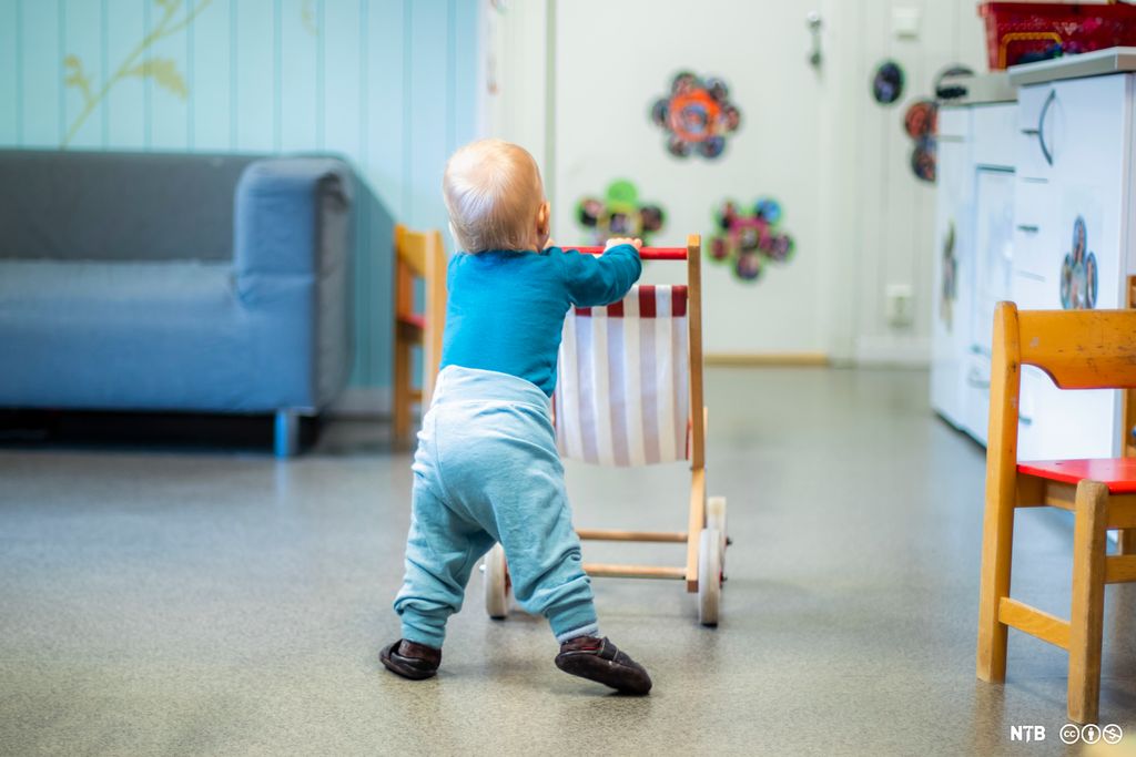 Et lite barn går ved å støtte seg til ei lita vogn. Foto.