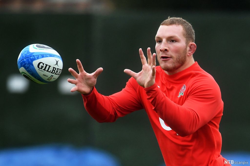 Mannleg rugbyspelar med raud drakt mot mørk bakgrunn. Står med åpne hender og ser konsentrert  mot ein blå og kvit rugbyball, klar til å ta imot. Foto.