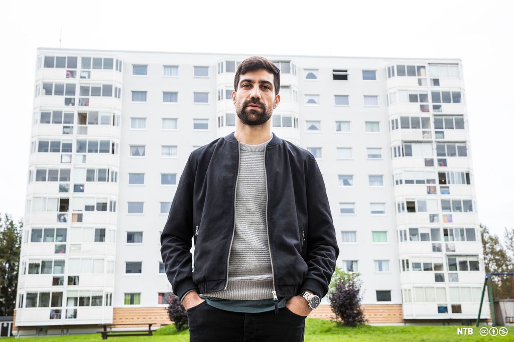 En ung mann med skjegg og svart jakke står foran ei høyblokk. Foto.