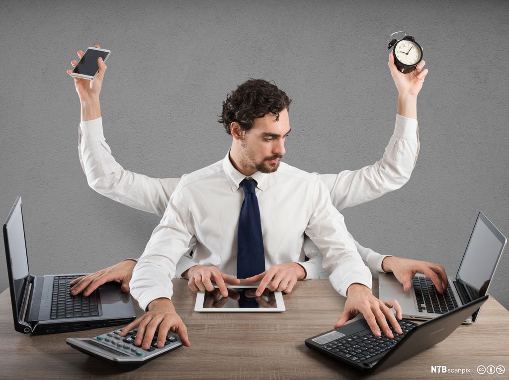 Stresset forretningsmann multitasker. Han har seks par armer. Hendene på armene gjør ulike ting. Fire av dem taster på ulike tastaturer, mens to av dem holder oppe henholdsvis en mobiltelefon og ei vekkerklokke. Mannen er kledd i hvit skjorte og slips. Han har mørkt, krøllete hår og kort skjegg. Foto.