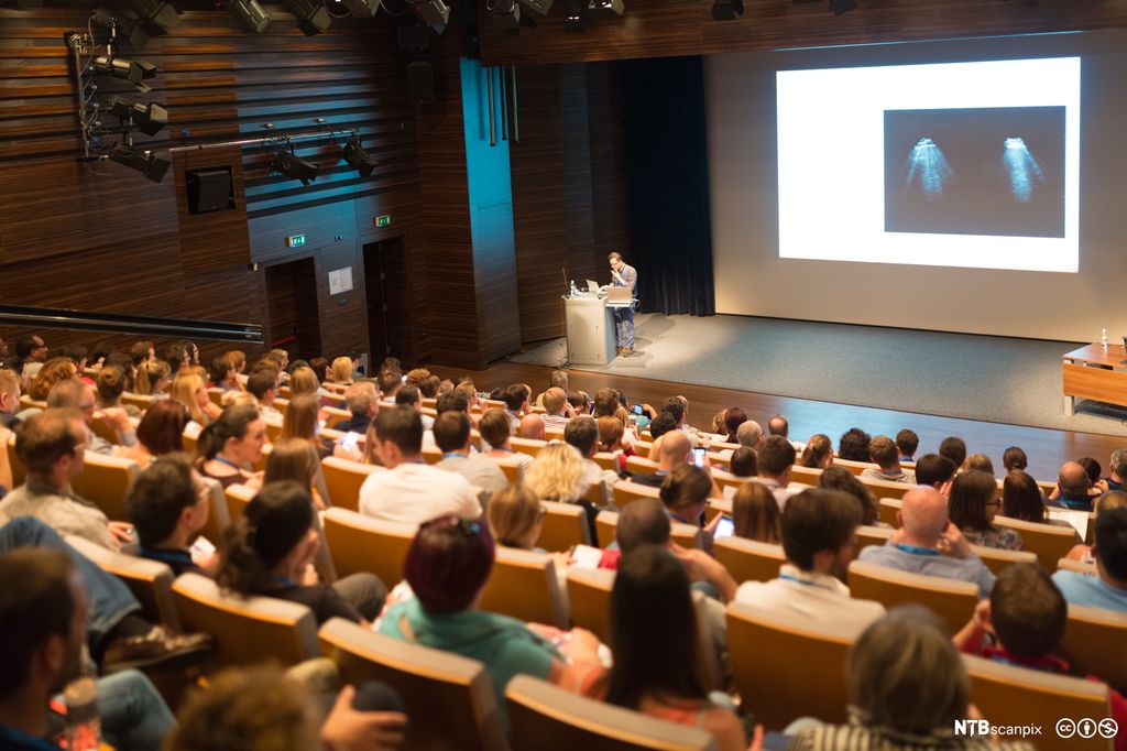Publikum på konferanse. Framme står en foredragsholder, og det er et bilde via videokanon på veggen bak. Foto.