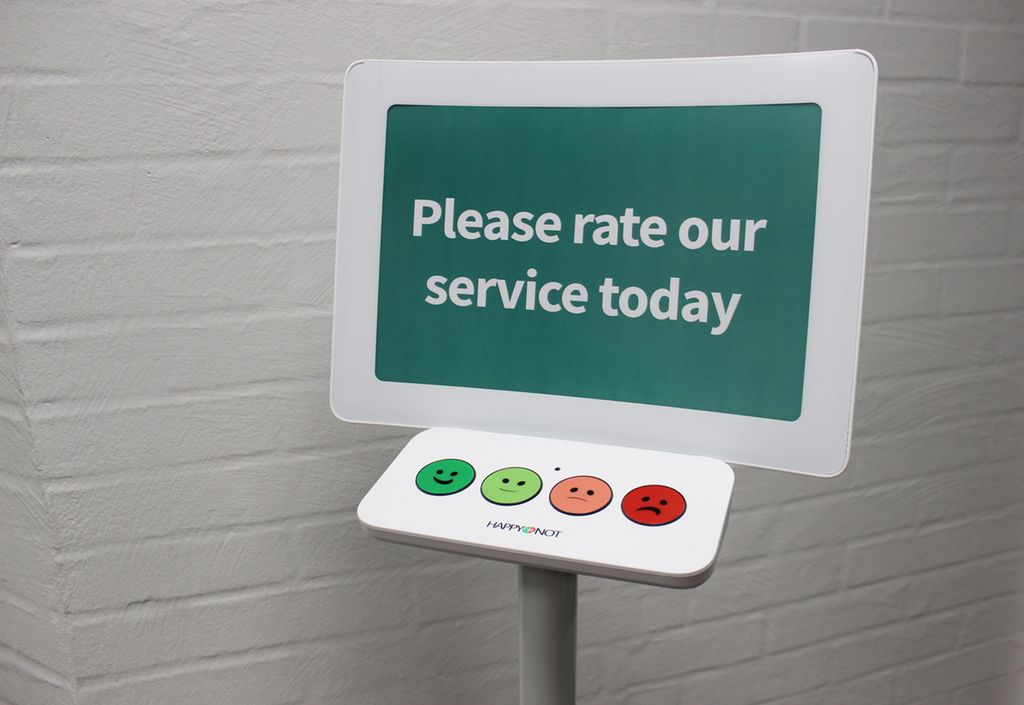 En skjerm med teksten Please rate our service today vises. Under skjermen er det fire knapper: en mørkegrønn med et smilefjes med et stort smil på, en lysegrønn med et smilefjes med et litt mindre smil på, en lyserød med et smilefjes med en liten sur munn på og en mørkerød med et smilefjes med en veldig sur munn på. Foto.  