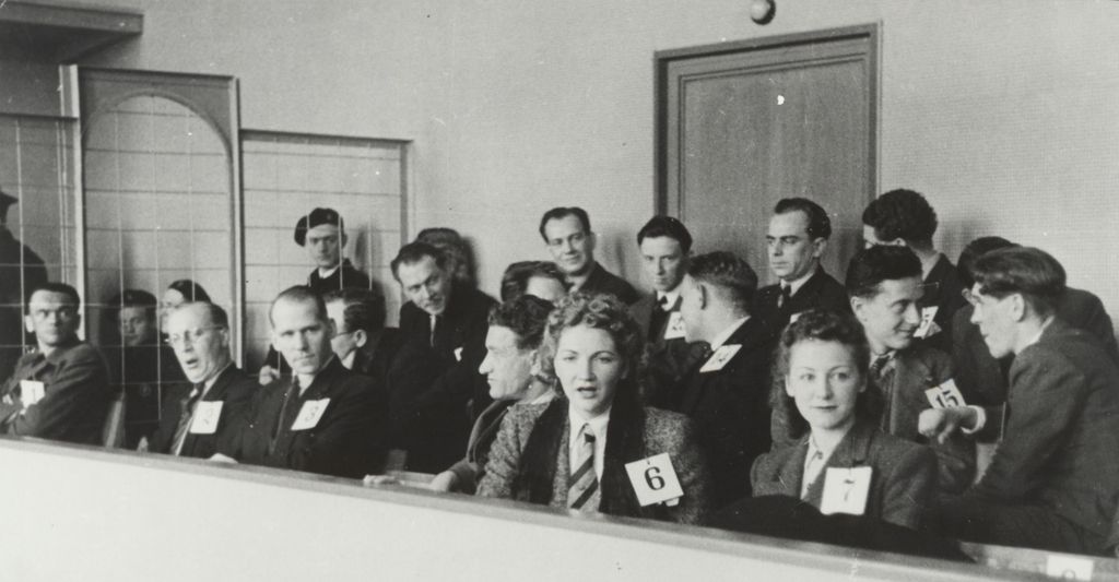 Medlemmene i Rinnanbanden sitter i tre rekker på tiltalebenkene i rettssalen. Det er to kvinner blant dem. Alle har nummerskilt på seg. Foto.