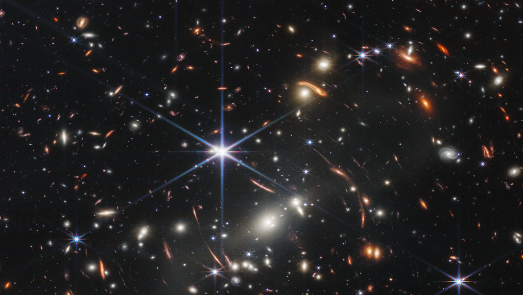 Tusenvis av små galaksar. Fargane deira varierer. Nokre er nyansar av oransje, mens andre er kvite. Dei fleste blir viste som uklare ovalar, men nokre få har distinkte spiralarmar. Framfor galaksane er det fleire stjerner. Dei fleste ser blå ut, og dei lyse stjernene har diffraksjonsspor som dannar eit åttepunkta stjerneform. Det er òg mange tynne, lange, oransje bogar som bøyer seg rundt sentrum av biletet. Foto.