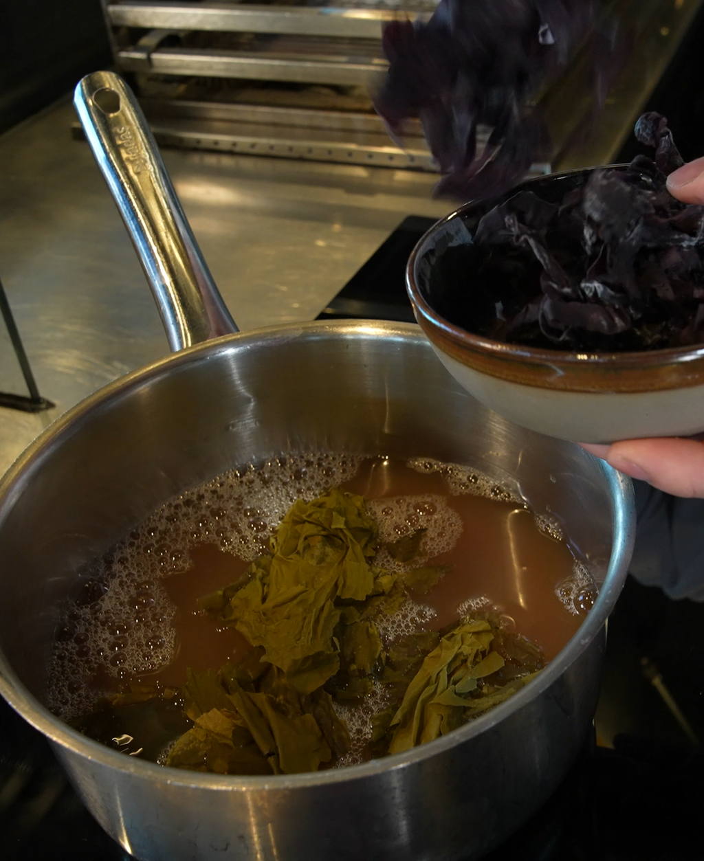 Ei gryte med fiskekraft. En kokk er i ferd med å tilsette tang og tare i gryten. Foto.