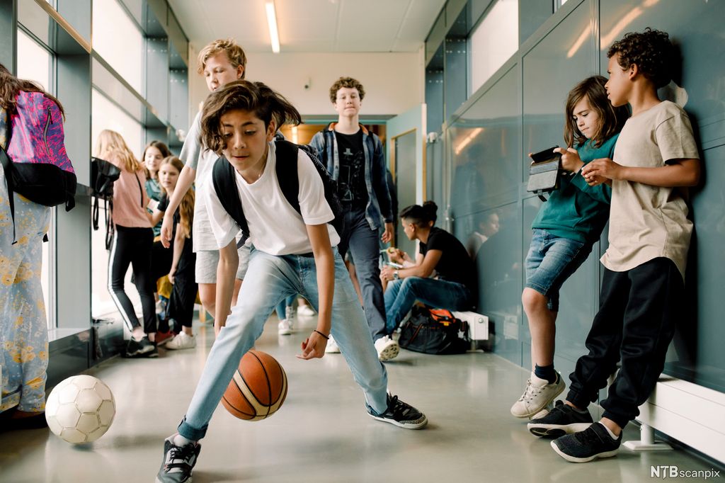Barn i ein skulekorridor. I midten ser vi ein gut som triksar med ein basketball. Langs veggane og bak guten står det andre barn. Nokon ser på ein iPad, nokon ser ut av vindauget. Foto.
