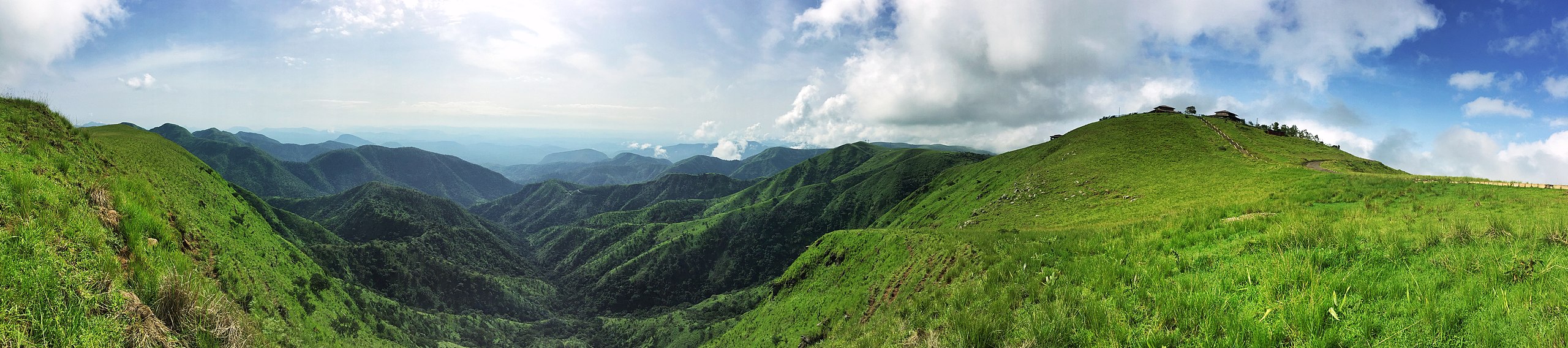 Photo: Landscape picture we see rows of green mountains disappearing into the distance. The sky above is blue, with a few clouds. 