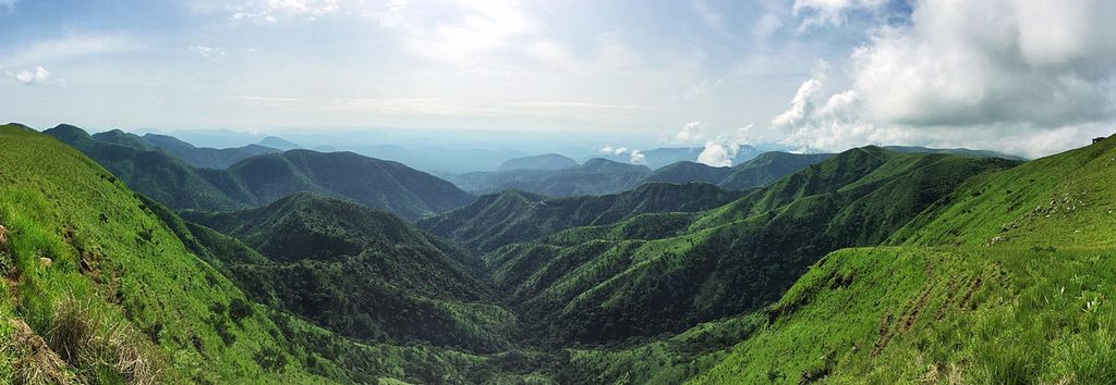 Photo: Landscape picture we see rows of green mountains disappearing into the distance. The sky above is blue, with a few clouds.