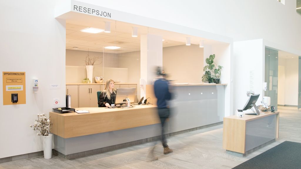 Foto av et resepsjonsområde der en ung kvinne sitter bak skranken og kikker opp på en person som går forbi.