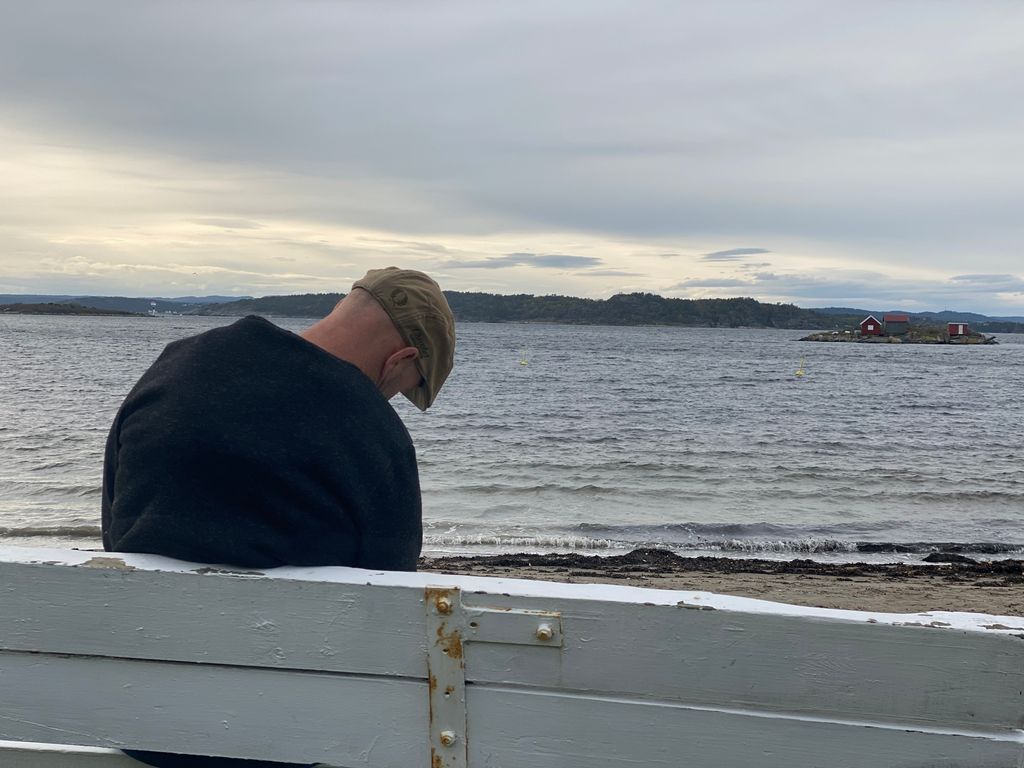 Mann som sitter på en benk på en strand. Hodet hans henger framover, og han sitter litt skjevt. Foto. 