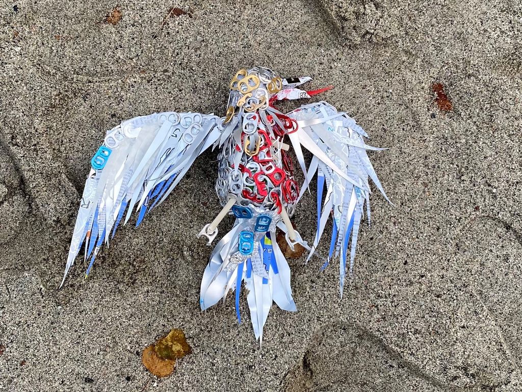 En fugl som er laget av deler fra metallbokser, ligger i sanden. Foto.